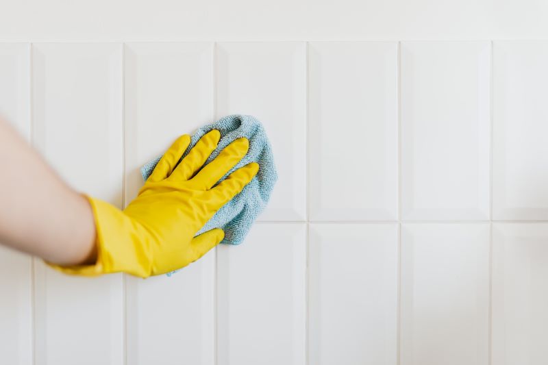 Woman’s arm with yellow rubber glove on hand cleaning white subway tile wall with blue cloth